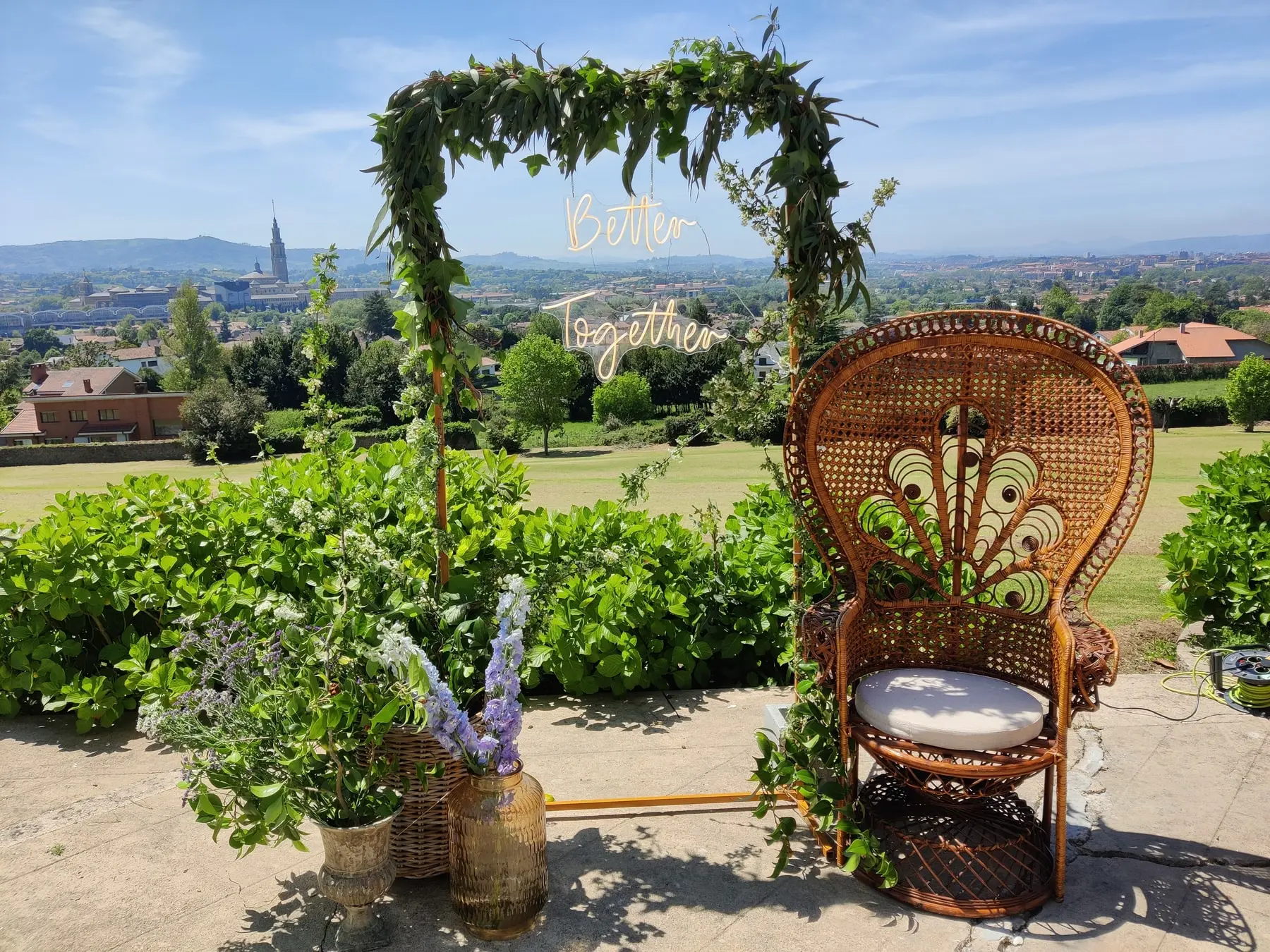 Un arco de flores y hojas con un sillón de mimbre, y Gijón al fondo bajo un cielo azul