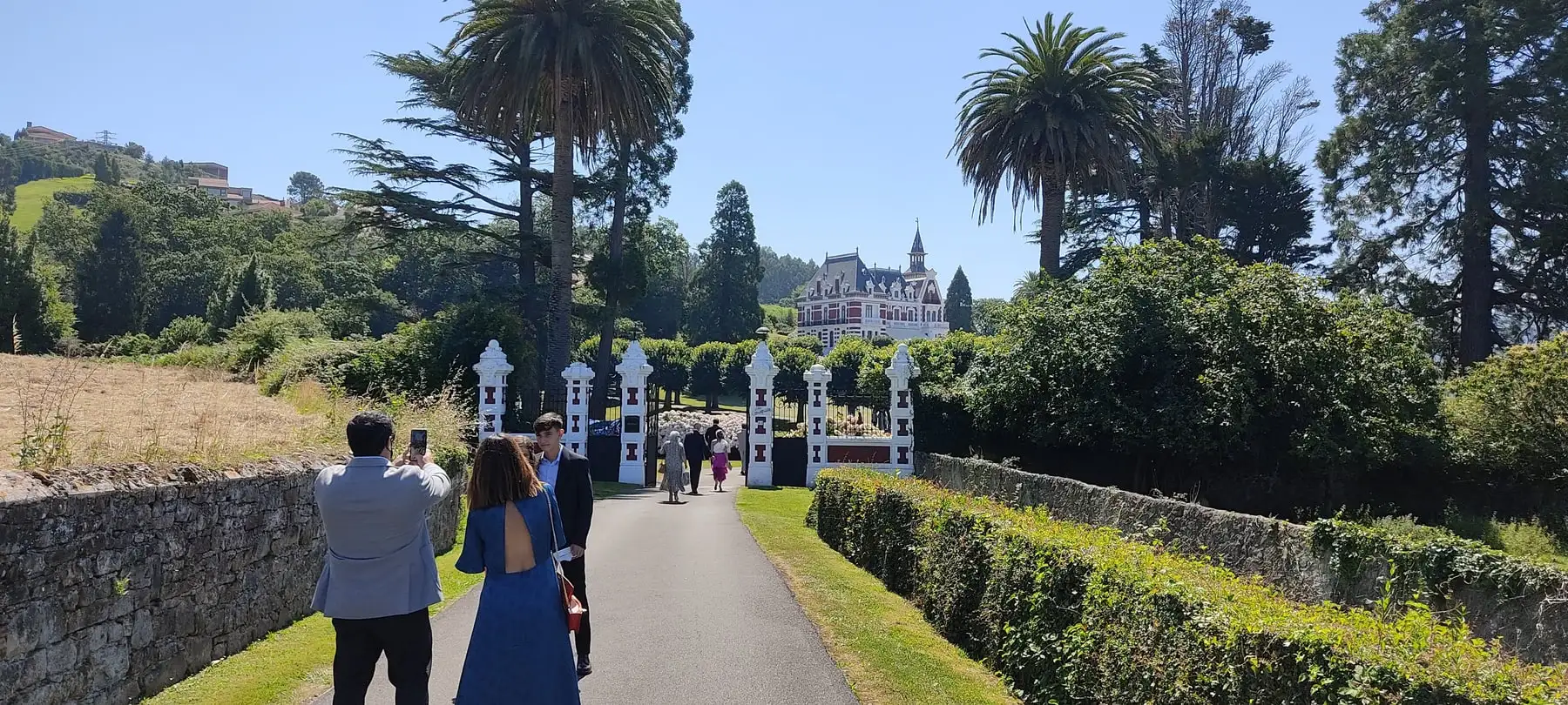 Un camino entre un muro de piedra y un seto, con invitados que caminan hacia el palacio y una verja blanca al fondo