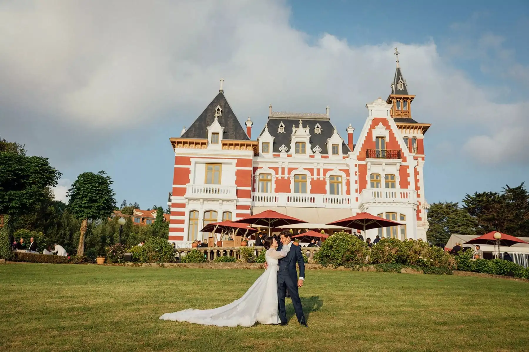 Los novios sobre el césped, delante del palacio, con las sombrillas rojas de la terraza detrás