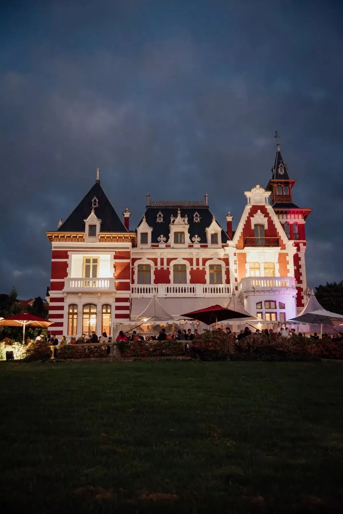 El palacio de noche, con las ventanas encendidas, los invitados bajo las sombrillas y un cielo azul oscuro