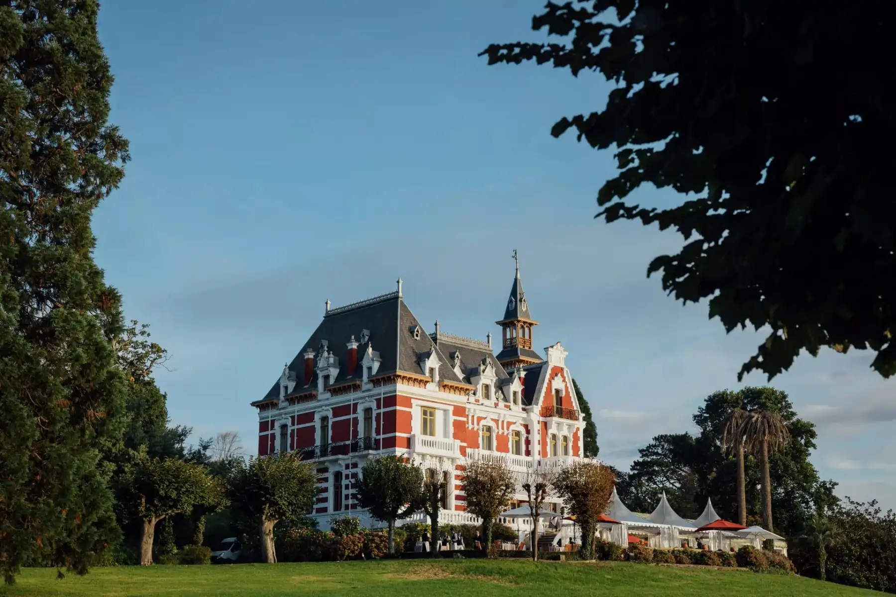 El palacio entre árboles a última hora de la tarde, con el sol dorando el ladrillo y las carpas blancas a la derecha