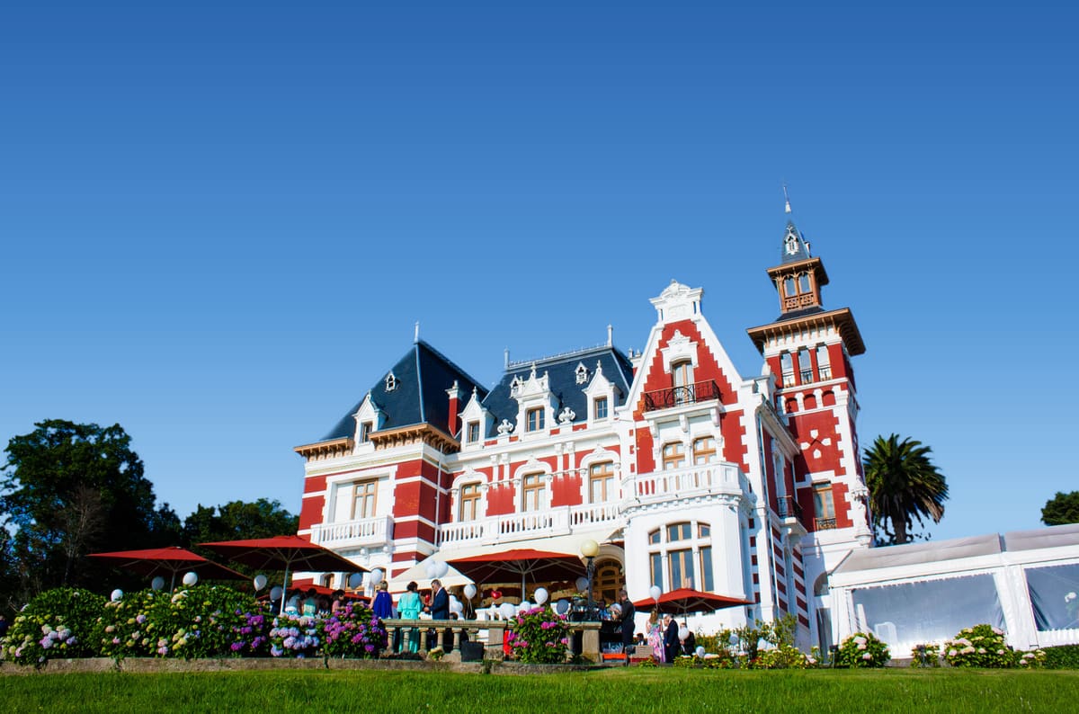 El Palacio de la Concepción visto desde el césped bajo un cielo azul: fachada de ladrillo rojo y piedra blanca, la torre a la derecha, sombrillas rojas y hortensias en la terraza